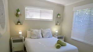Un dormitorio ordenado con una cama grande, dos mesitas de noche con lámparas, una ventana con persianas y estantes en la pared con plantas.