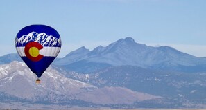 Un globo aerostático con diseño de montaña y bandera, volando sobre un paisaje montañoso.