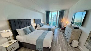 A hotel room with two beds, a desk, a dresser, a TV, and a view of the city. - Balcony Ocean Views | Pool, Gym, Wi-Fi & Parking (Miami)