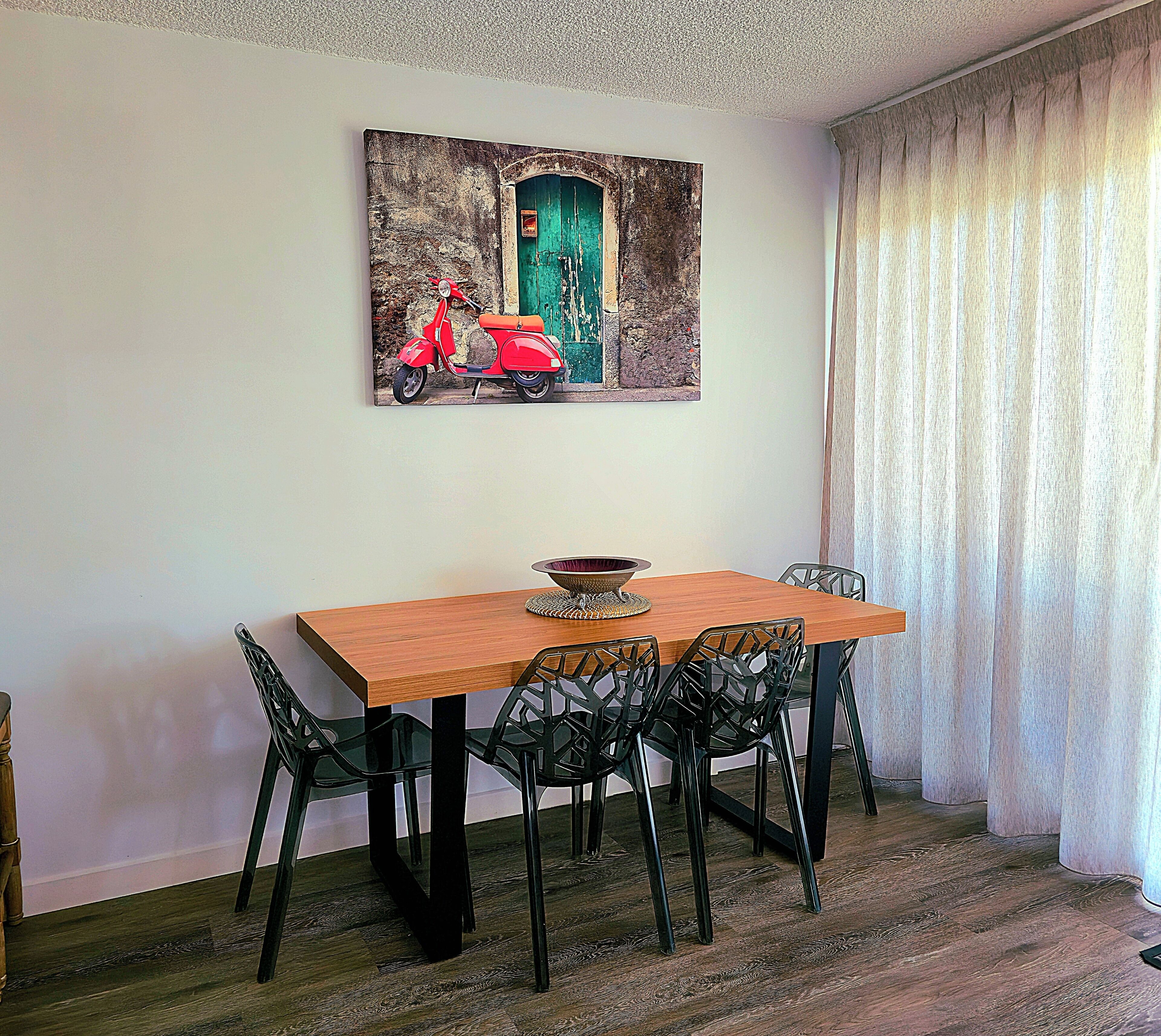 A dining area with a wooden table, black chairs, and a painting of a red scooter on the wall.
