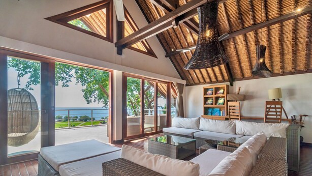 A spacious living area with a large sofa, a glass coffee table, and wicker furniture. There is a bookshelf with books and decorative items, and a hanging light fixture. The room has large windows with a view of the outdoors. - Luxury Beach & Cliff Villa with Unbelievable Panoramic Views.  (Seririt)
