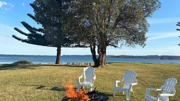 A grassy area with a fire pit, three white chairs, and a large tree.