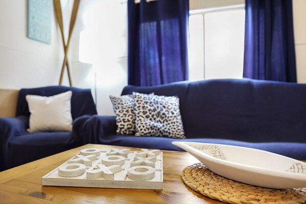 Una sala de estar con un sofá azul oscuro, cojines estampados, una mesa de centro de madera con un juego de mesa y un mantel tejido.