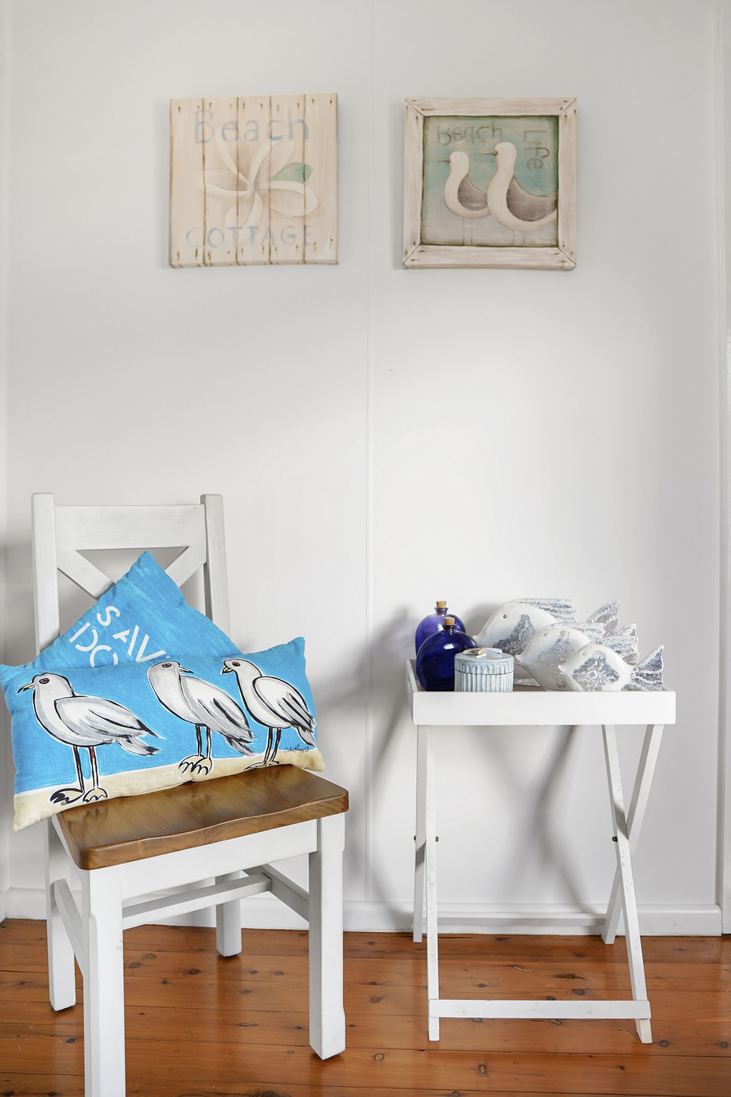 Een witte stoel met een blauw kussen waarop meeuwen afgebeeld zijn, een klein wit bijzettafeltje met decoratieve objecten, en twee ingelijste kunstwerken aan de muur.