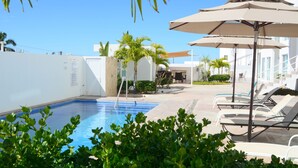 A pool area with lounge chairs and umbrellas, surrounded by greenery and a white building. - Spectacular Master Penthouse - Best Ocean view and sunsets on the Sea of Cortez (La Paz)