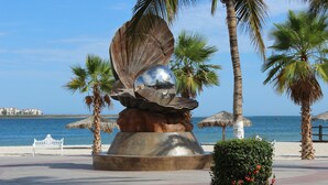 A sculpture of a shell with a reflective sphere, palm trees, a bench, and a beach view. - Spectacular Master Penthouse - Best Ocean view and sunsets on the Sea of Cortez (La Paz)