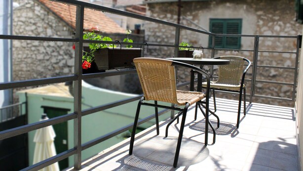 Un balcon agrémenté de chaises et d’une table en osier, d’où l’on peut admirer un bâtiment en pierre surmonté d’un toit en tuiles.