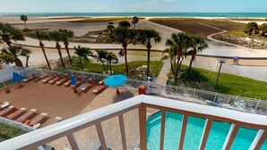 A pool area with lounge chairs and umbrellas, overlooking a beach with palm trees and a clear sky.