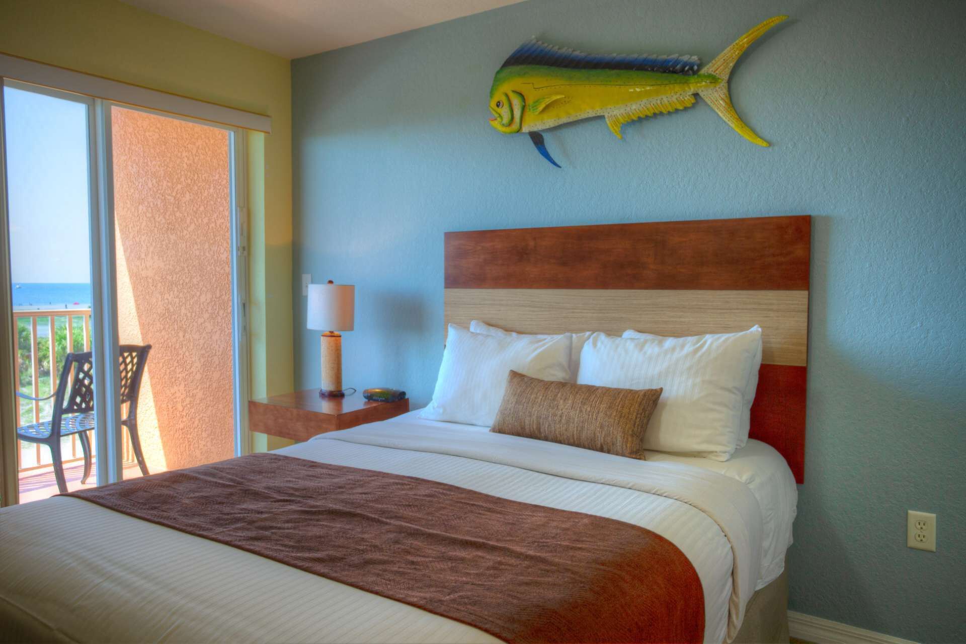 A hotel room with a bed, a nightstand, a lamp, a chair, and a large fish decoration on the wall.