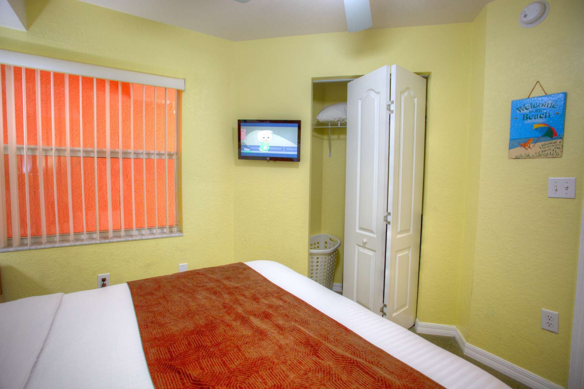 Een slaapkamer met een bed, een televisie aan de muur, een raam met oranje luiken en een deur die naar een andere kamer leidt.
