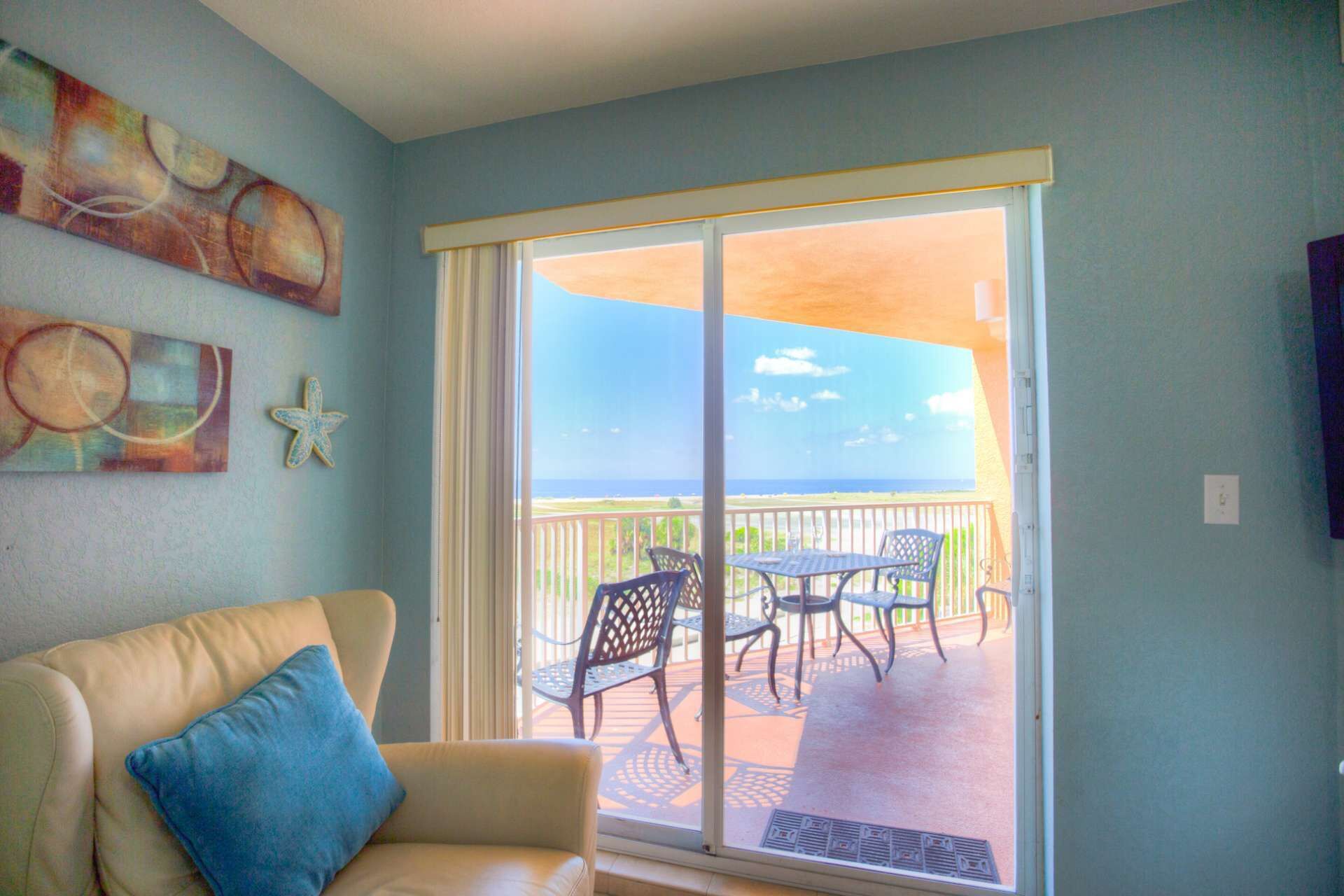 Una habitación con un sofá color beige, una almohada azul, una decoración con forma de estrella de mar y vistas al océano a través de una puerta corrediza de vidrio.