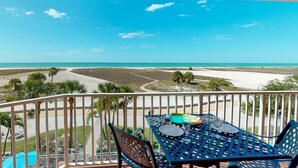 A balcony with a table and chairs, overlooking a beach with a clear view of the ocean and distant shoreline.