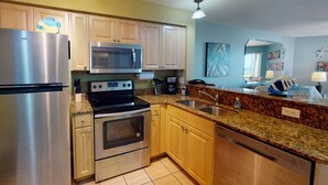 A modern kitchen with stainless steel appliances, wooden cabinets, granite countertops, and a view of the living area.