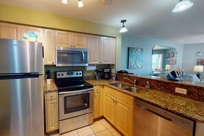 A modern kitchen with stainless steel appliances, wooden cabinets, granite countertops, and a view of the living area.