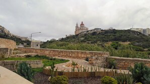 Un chemin de pierre mène à un bâtiment historique surmonté d’un dôme rouge, entouré d’une végétation luxuriante et d’un mur de pierre.