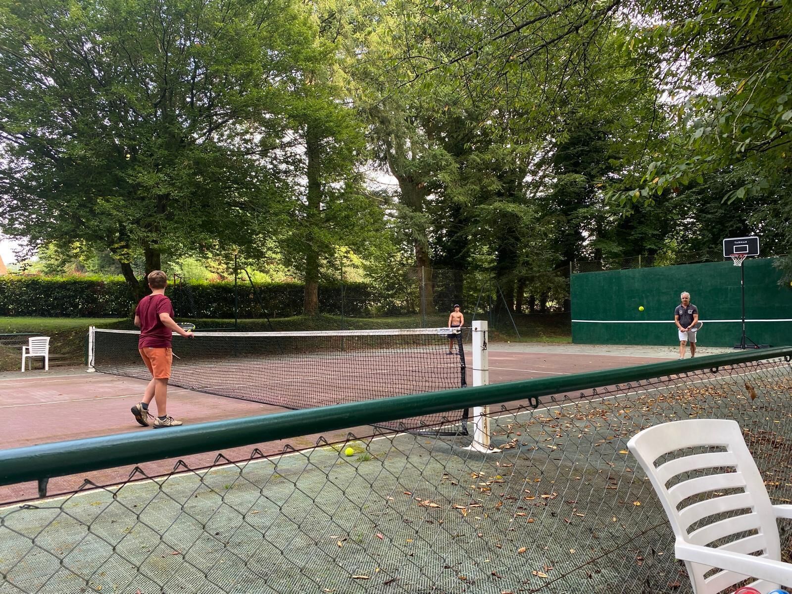 Un campo da tennis all'aperto con una rete, circondato da alberi e un canestro da basket.