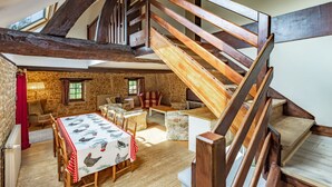 Un interior rústico con una escalera de madera, paredes de piedra, una zona de comedor con una mesa preparada para cuatro personas y una acogedora disposición de asientos.