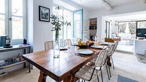 A dining area with a wooden table set for six, surrounded by wicker chairs. A large window provides natural light, and a plant is placed in the center.