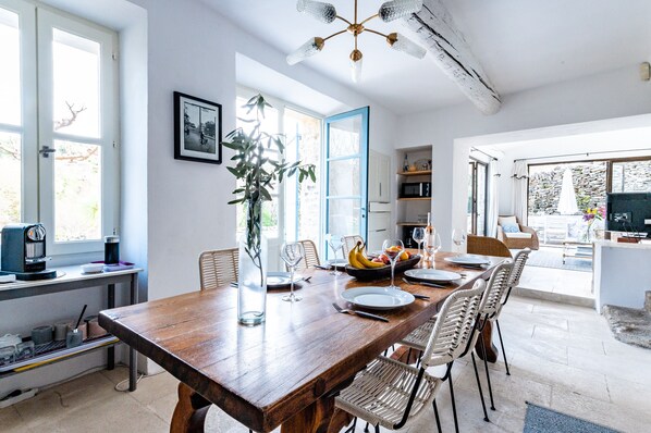 A dining area with a wooden table set for six, surrounded by wicker chairs. A large window provides natural light, and a plant is placed in the center.