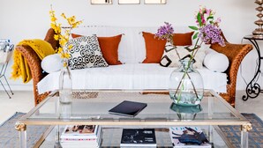 A living room with a white sofa, patterned cushions, a glass coffee table with magazines, and a vase with flowers.
