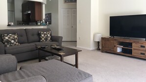 A living room with a grey sofa, a coffee table, a flat-screen TV on a wooden stand, and a kitchen area in the background. - Premier Host Rated Villa - Private solar heated pool & family games room   (Davenport)