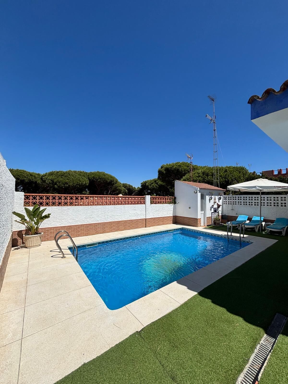 Una piscina con agua azul cristalina, rodeada por un muro blanco con detalle de ladrillo rojo y un área de césped verde con sillas de descanso.