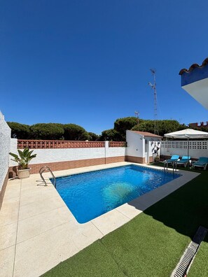 A swimming pool with a clear blue water, surrounded by a white wall with a red brick border, and a green lawn area with lounge chairs. - Ana's House. Villa on Mazagón Beach with parking and a private pool. (Moguer)