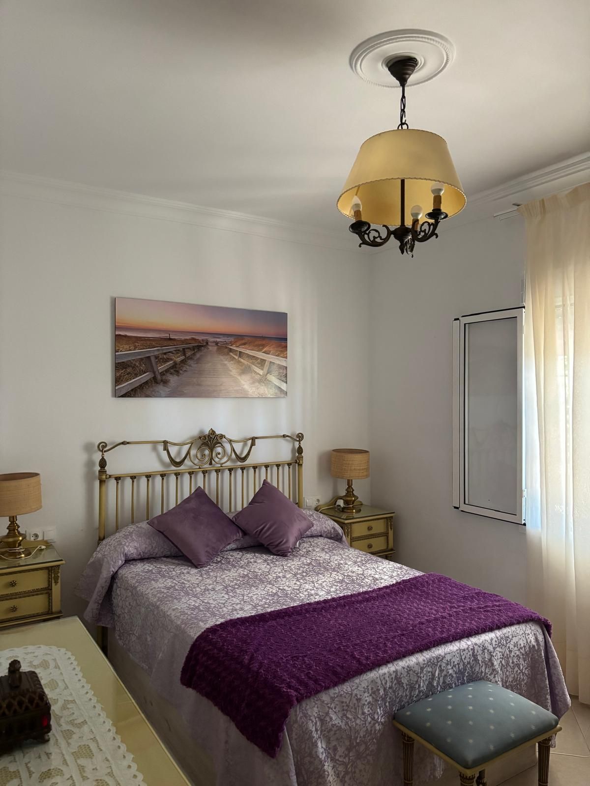 Un dormitorio con cama, mesita de noche, lámpara, un cuadro en la pared y ventana con cortinas.