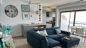 A modern living room with a blue sofa, a small white table, and a kitchen area in the background. - The Moll Apartment - Seaside Holiday Home (Il-Mellieha)
