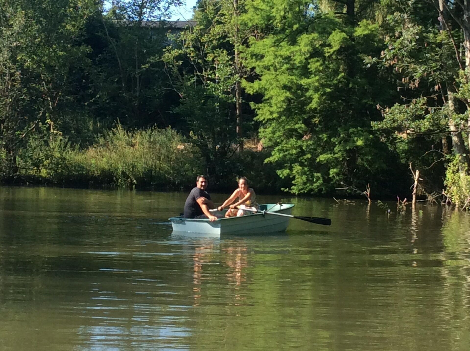 Two people rowing a small boat on a calm lake surrounded by dense trees.