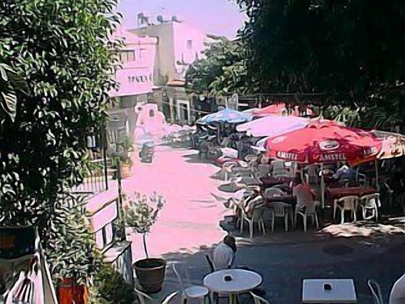 Un café al aire libre con mesas, sillas y sombrillas, con una calle al fondo.