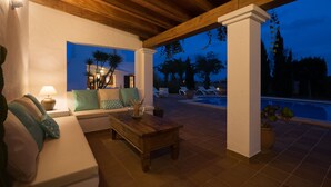 Ein Außenbereich mit Terrasse, Holzboden, weißen Wänden, einer Couch mit Kissen, einem Couchtisch und einem Pool im Hintergrund.