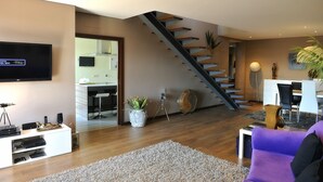 Een moderne woonkamer met een trap, een flatscreen televisie, een eethoek en een keuken, zichtbaar door een open deur.