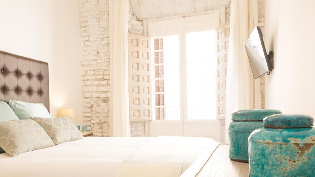 A bedroom with a bed, a stone wall, a window with shutters, and a TV mounted on the wall.