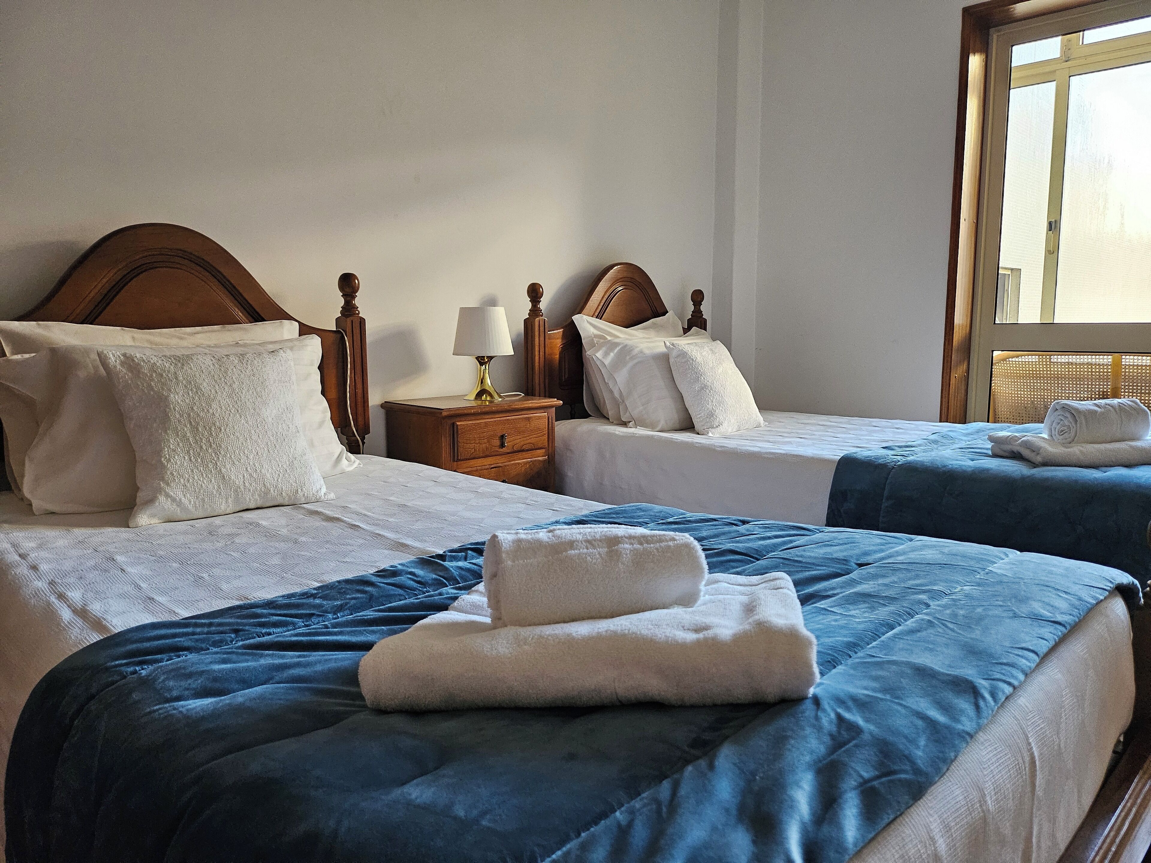 Une chambre d’hôtel avec deux lits, chacun doté d’une tête de lit en bois, de draps blancs et d’une couverture bleue. Une table de chevet avec une lampe se trouve entre les deux lits.