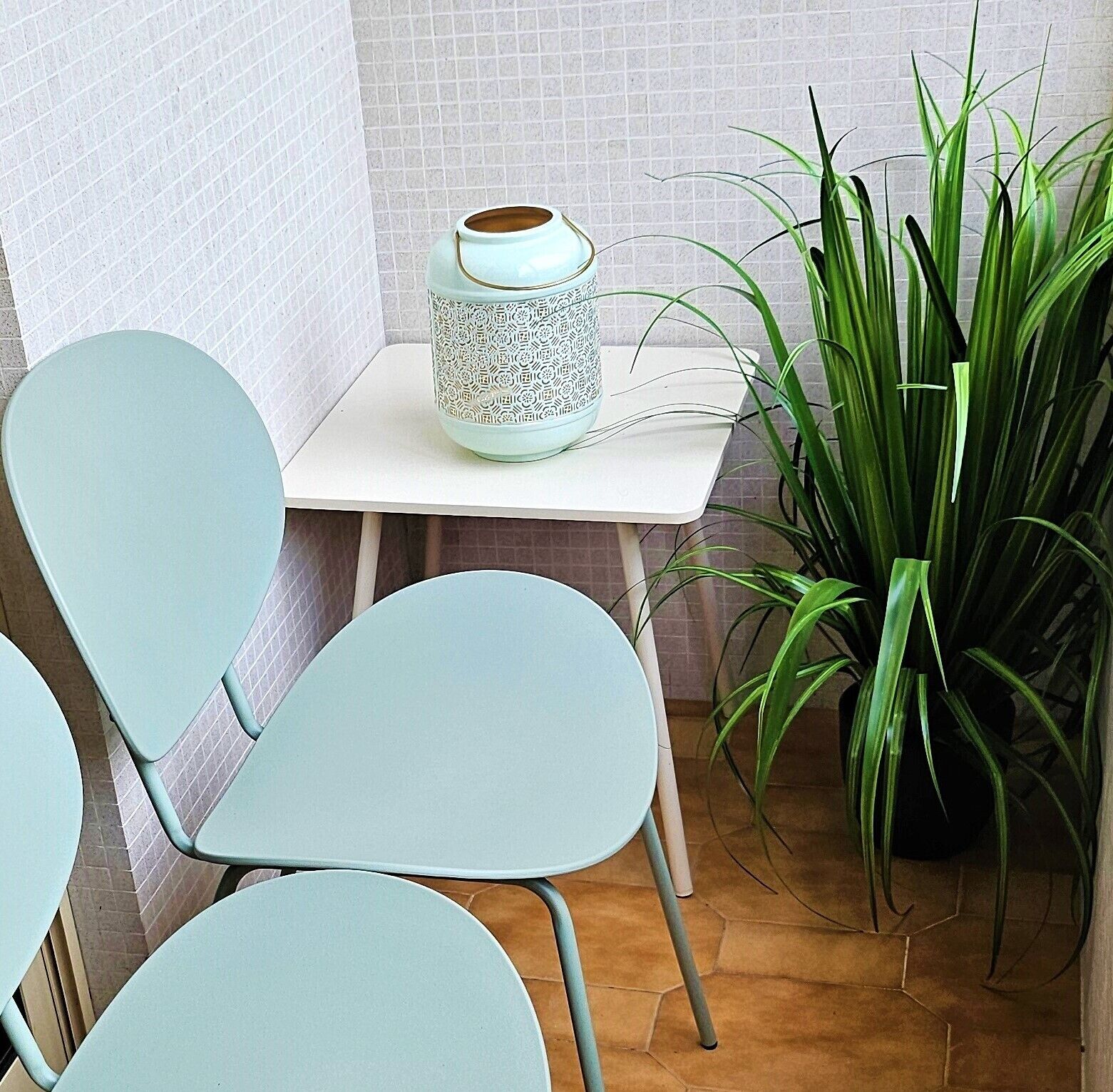 Een hoek met een witte betegelde muur, een witte tafel met een vaas met dessin, een groene plant en twee lichtblauwe stoelen.