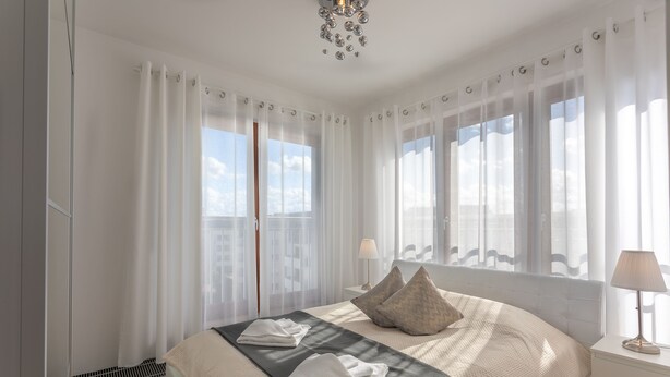 Une chambre à coucher moderne avec un grand lit, des rideaux blancs et une vue sur le ciel par les fenêtres.