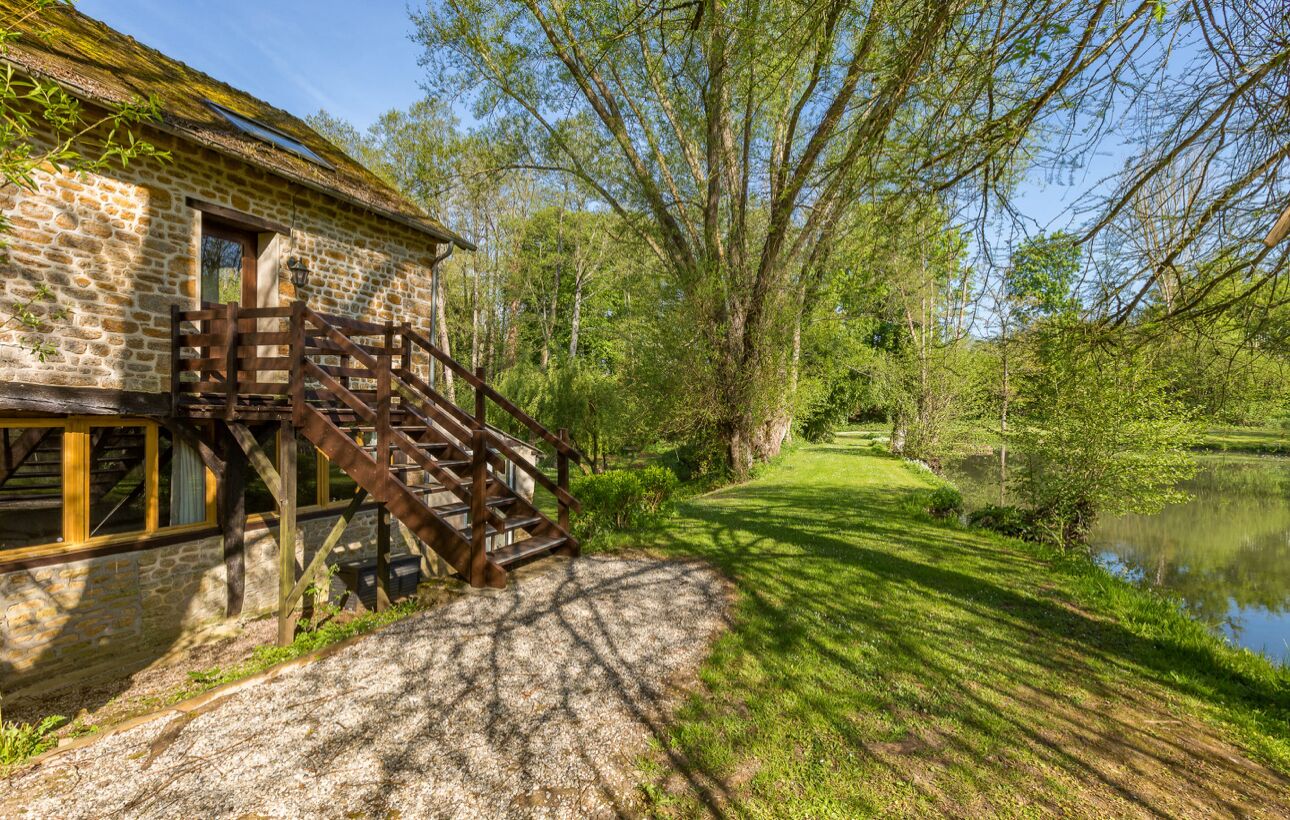 Un bâtiment rustique en pierre, doté d’un escalier en bois menant à une fenêtre, est entouré d’une végétation luxuriante et d’un étang.