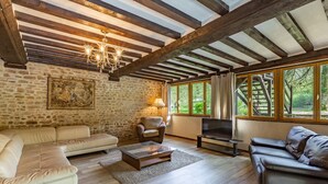 A cozy living room with stone walls, wooden beams, a fireplace, and large windows offering a view of the outdoors.