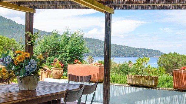 A wooden table with chairs under a pergola, overlooking a pool and a scenic view of a lake and hills. - Detached villa 50 meters from the sea (Isola d'Elba)