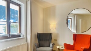 Ein Zimmer mit grauem Sessel, rotem Sessel, rundem Spiegel, kleinem Tisch mit Buch und Fenster mit Vorhängen.