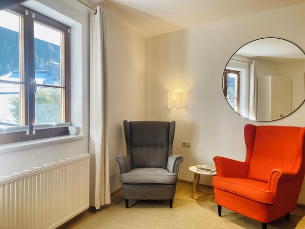 A room with a grey armchair, a red armchair, a round mirror, a small table with a book, and a window with curtains.