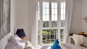 Une chambre avec une grande fenêtre, un lit avec une literie à rayures, une lampe et une vue sur la verdure à l’extérieur.