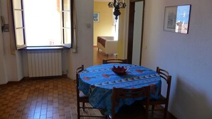 Un coin repas avec une table ronde recouverte d’une nappe à motifs floraux, entourée de chaises en bois. Un lustre est suspendu au-dessus de la table, et une fenêtre avec des rideaux est visible à l’arrière-plan.