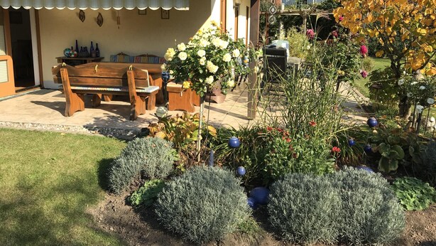 A patio with a wooden bench and table, surrounded by a garden with various plants and flowers. - Comfortably furnished apartment in Pastetten district Erding (Pastetten)