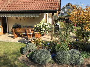 A patio with a wooden bench and table, surrounded by a garden with various plants and flowers. - Comfortably furnished apartment in Pastetten district Erding (Pastetten)