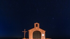 Eine kleine, abgelegene Kirche mit Kreuz und Glockenturm, umgeben von einer felsigen Landschaft unter einem sternenübersäten Nachthimmel.