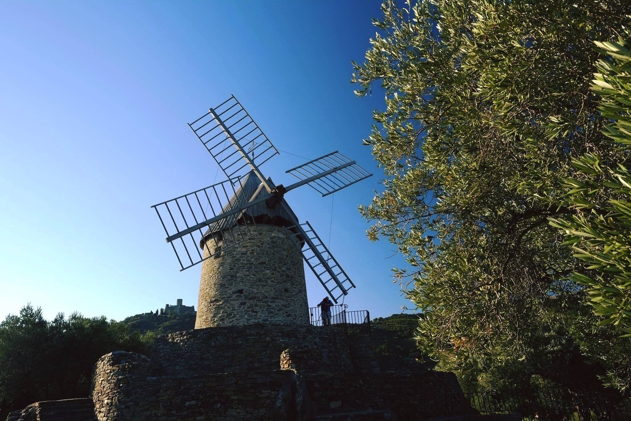 A windmill with a stone base and metal blades, surrounded by trees and a clear sky.