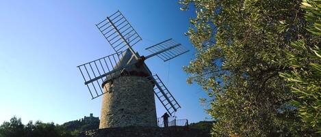 A windmill with a stone base and metal blades, surrounded by trees and a clear sky.