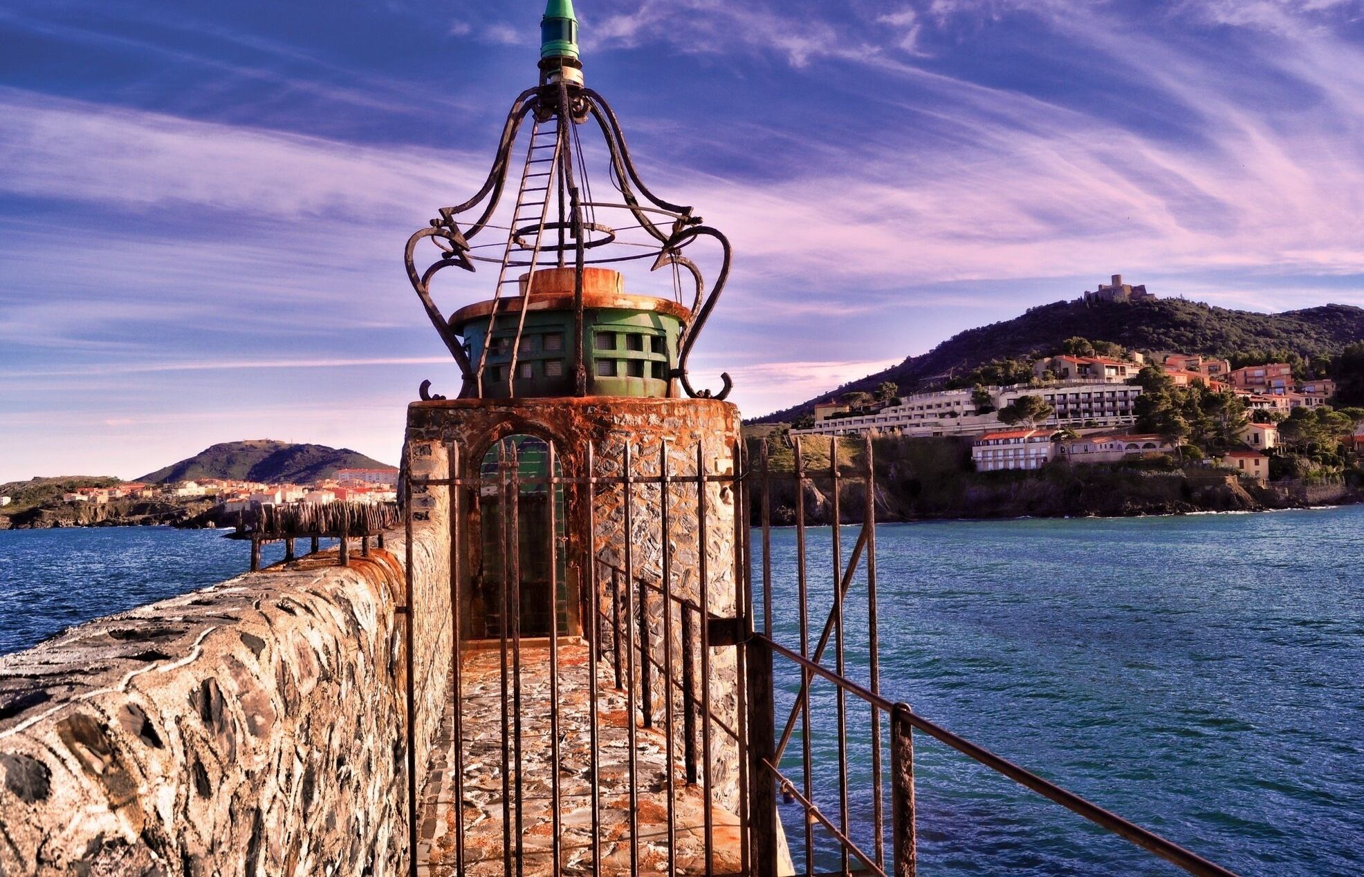 A coastal lighthouse with a stone base and metal structure, overlooking a body of water with a town in the background.